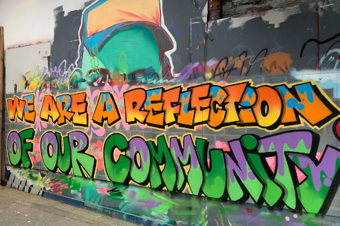 Graffiti mural reads ‘WE ARE A REFLECTION OF OUR COMMUNITY’ in bold orange, green, and purple letters on a colourful wall, linked to Cardiff-based non-profit social enterprise Media Academy